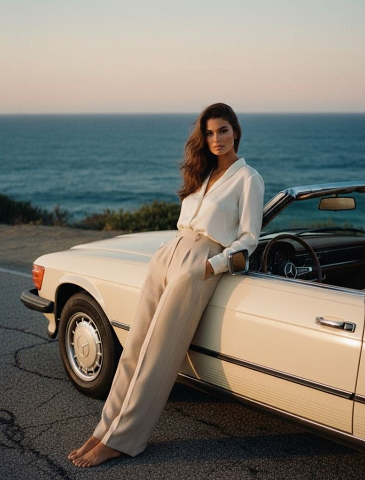 A woman in cream tailoring leaning on a vintage convertible by the sea at dusk